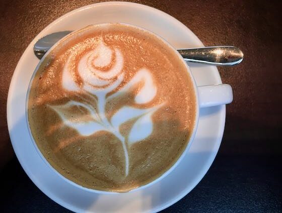 Eine Rose mit Latte Art gezeichnet, also der Kunst mit perfekten Milchschaum ein Bild in den Kaffee zu malen. Horst hat dies bereits auf Meisterschaften getan. Auf dem Bild sieht man die weiße Tasse von oben, auf einer Untertasse mit einem Cappuccino Löffel an der oberen Seite