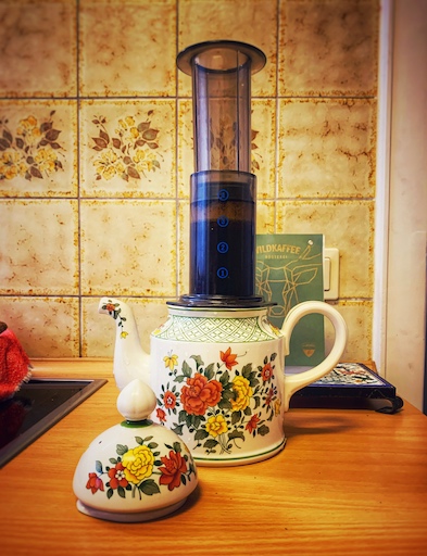 AeroPress Coffee and Espresso Maker, das Gerät steht ganz unüblich auf einer mit Blumen bedruckten alten und ganz klassischen Kaffeekanne, in einer alten Küche. Spannende Kombination aus Moderne und Tradition.