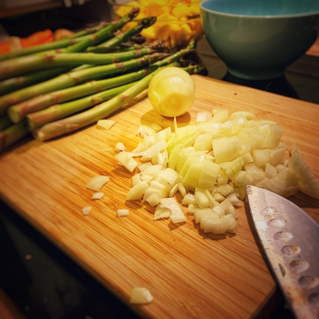 Mit Gleichmäßigkeit geschnittene Zwiebel, bereit zum Kochen, liegt auf einem Holzbrett. Im Vordergrund sieht man noch die Messerspitze und im Hintergrund den grünen Spargel.