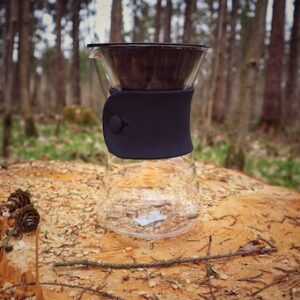 Der gläserne Hario V60 Drip Decanter steht dekorativ auf einem Holzstamm im Wald. Die Kanne an sich ist gläsern, der Silikonkragen sowie der Filterhalter sind schwarz.