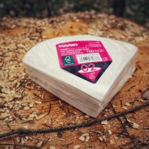 Hario V60 Filterpapier 02 aus EU Produktion. Eine Packung mit 100 Stück liegt dekorativ auf einem Holzstamm im Wald.