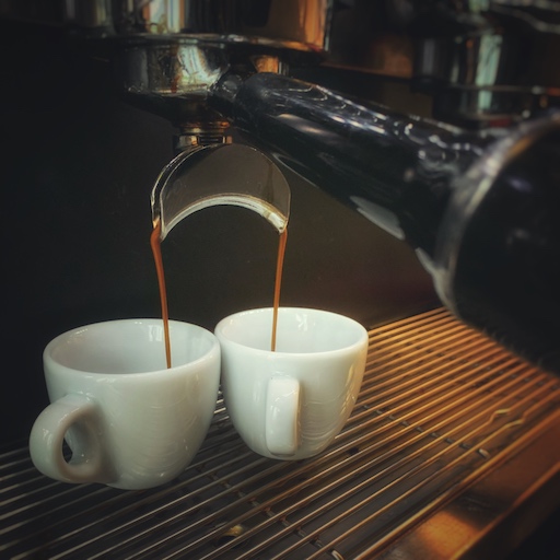 Zwei Espresso Tassen auf dem Abtropfgitter einer schwarzen Siebträgermaschine. Der Kaffee läuft aus den Silbernen Auslässen. Einfach besseren Espresso machen weiht dich in die Geheimnisse dieses Bildes ein. Ein Barista at Home Kurs online und offline möglich.