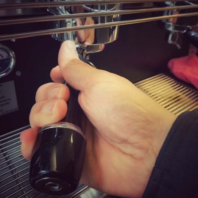 In Nahaufnahme sieht man eine Hand, welche einen Siebträger in eine professionelle Kaffeemaschine einhängt. Dies ist fast immer Bestandteil des Workflows an Kaffeebars.