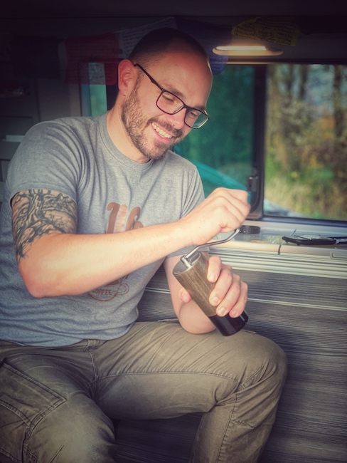 Horst von einfach mal Kaffee beim Kaffee machen im Camper. Er sitzt in einem Campern und mahlt mit der Handmühle frisch den Kaffee. Man sieht, dass es ihm spaß macht.