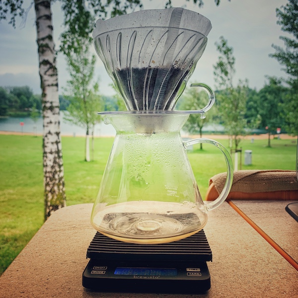 Es gibt viele Wege guten Kaffee im Camper zu machen. Hier sehen wir einen Hario V60 Filter in der Küche eines Vans. Es wird gerade Kaffee darin gebrüht. Im Hintergrund viel Grün, eine Buche und ein See.