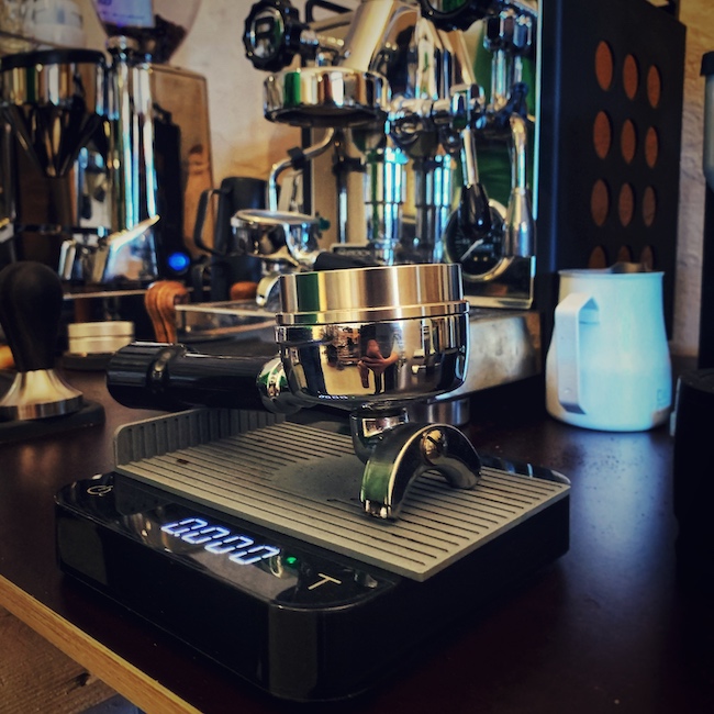 Ein Siebträger einer Rocket Espressomaschine steht auf einer Acaia Luna Kaffee Waage. Im Hintergrund sieht man die Maschine, sowie eine Mühle und Zubehör stehen. Alles samt ist nah aufgenommen und befindet sich auf einer schwarzen Barista Bar.