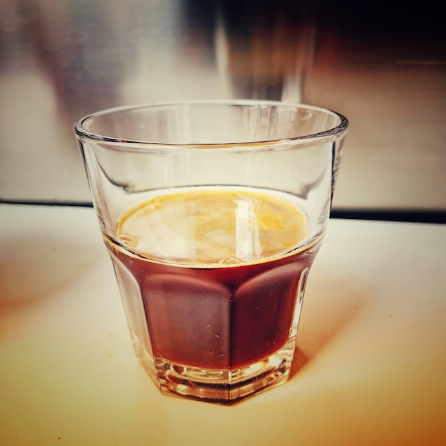 Ein Glas gefüllt mit Kaffee Americano auf einer weißen Bar vor einer silbernen Siebträgermaschine. Der Americano ist Nussbraun, seine Crema goldgelb.