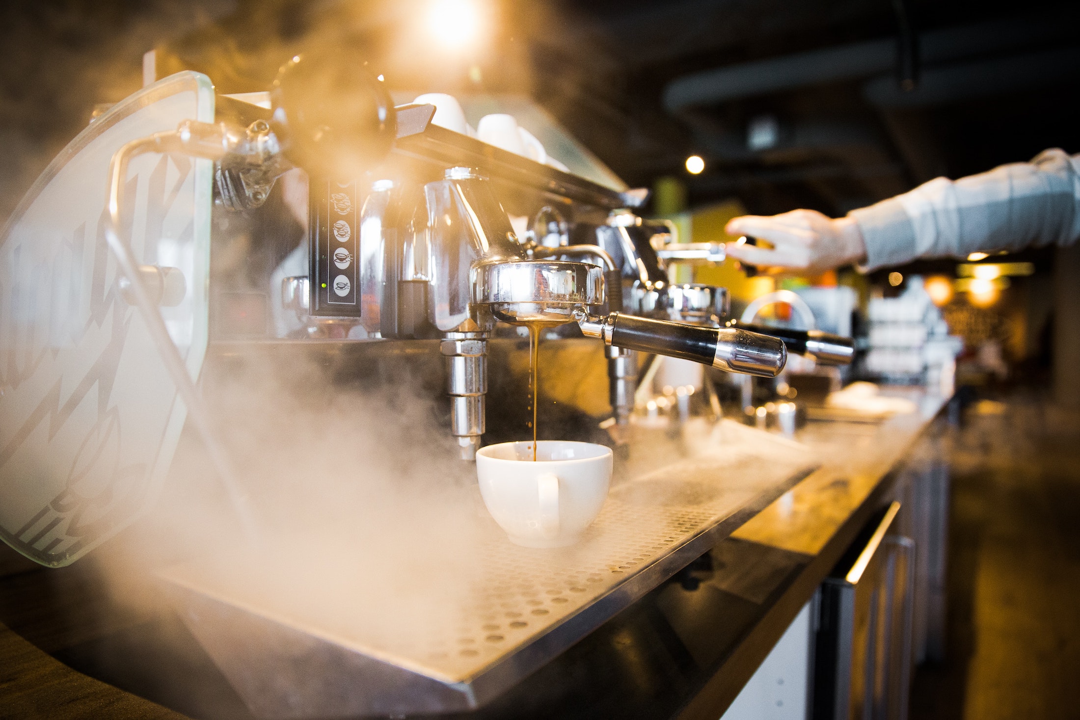 Eine silberne Siebträgermaschine steht auf der Bar und in ihrem eigenen Dampf. Der Barista an dieser Kaffee Bar hält einen Hebel auf der anderen Seite gedrückt.