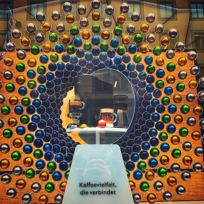 Ein dekorativer Kreis in Art eines Pfauenschwanzes aus Kaffee Kapseln, um eine Kapsel Kaffee Maschine angereiht in einem Schaufenster. Auf dem Sockel steht Kaffeevielfalt die Verbindet.