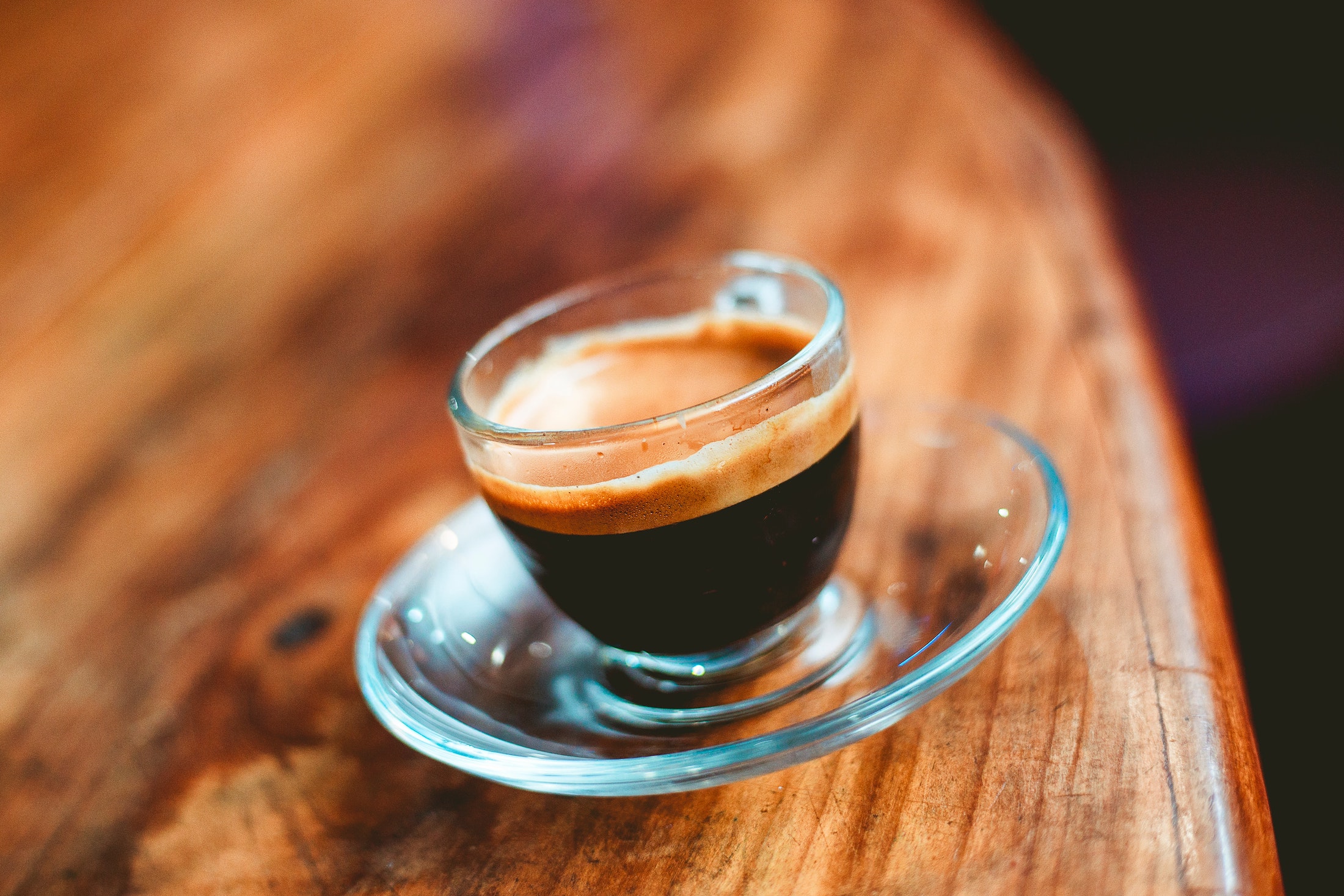 Eine gläserne Espresso Tasse auf einem Holztisch. Man erkennt gut die dunkle Kaffee Flüssigkeit und die nussbraune Crema auf dem Kaffee.