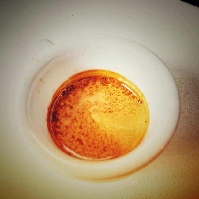Eine weiße Espresso Tasse aus weißem Grund, gefüllt mit nussbrauner Crema, die einige Tierflecken aufweist.