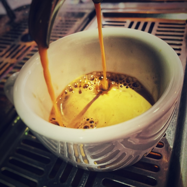 Eine weiße Tasse steht auf dem Abtropfgitter einer Espressomaschine. Gerade fließt goldgelber Espresso aus einem doppelten Siebträger in die Tasse und die Crema entsteht auf dem Kaffee.