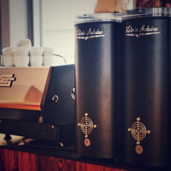 Zwei große schwarze Mythos One Kaffeemühlen stehen auf einer Barista Bar direkt neben einer La Marzocco GS3 Siebträgermaschine.