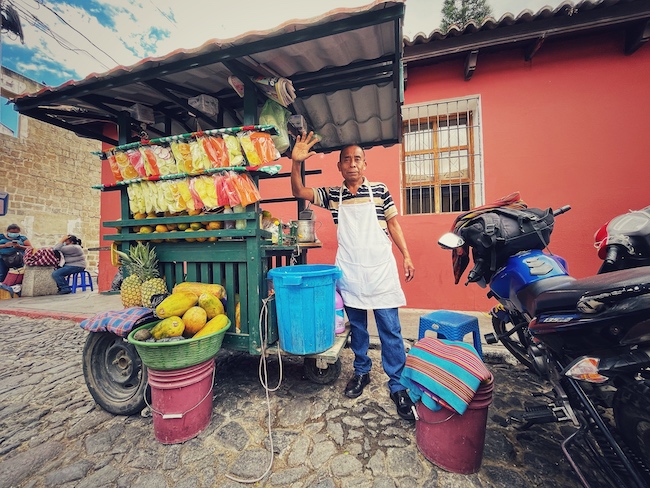 Fruchtverkäufer an seinem Stand vor einem bunten Haus in Antigua Guatemala.