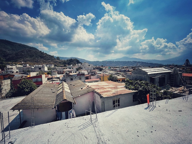 Blick über die beton Bauten und Dächer von Jocotenango. Im Hintergrund sieht man einen Vulkan.