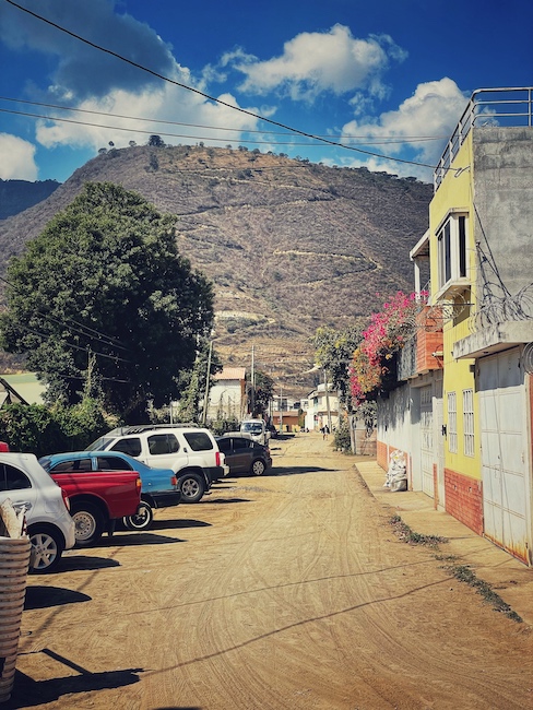 Eine Straße in Jocotenango. Am Rand stehen ein paar Autos, die Straße ist eine Staubpiste, im Hintergrund ist ein Berg.