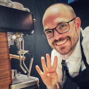 Messe Barista Horst von einfach mal Kaffee winkt in die Kamera, während er an der Espressomaschine steht.